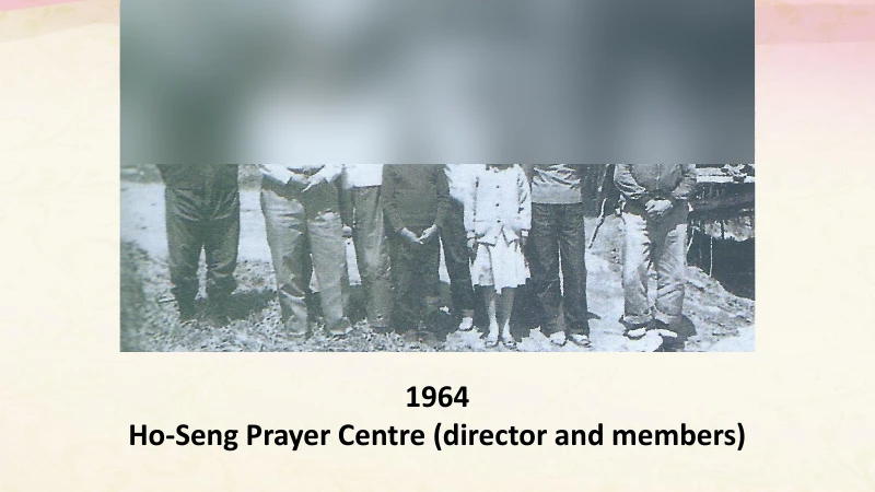 Training-deck page with a black-and-white group photo of about a dozen adults and children standing outside a hut, faces blurred, captioned 1964, Ho-Seng Prayer Centre (director and members).