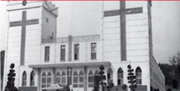 Black-and-white photo of the front of a white church building with a large cross on each of its two towers and arched windows on the ground floor.