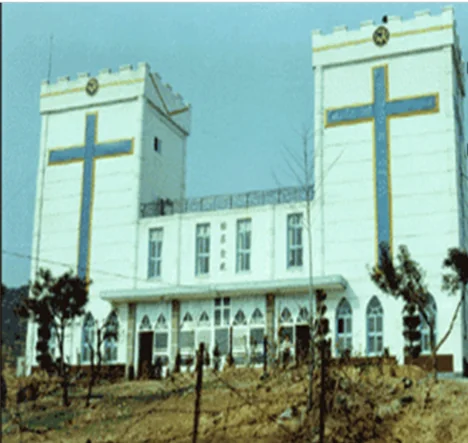 Colour photo of the front of a white church building with two crenellated towers, each bearing a large blue cross, with bare ground and young trees in front.
